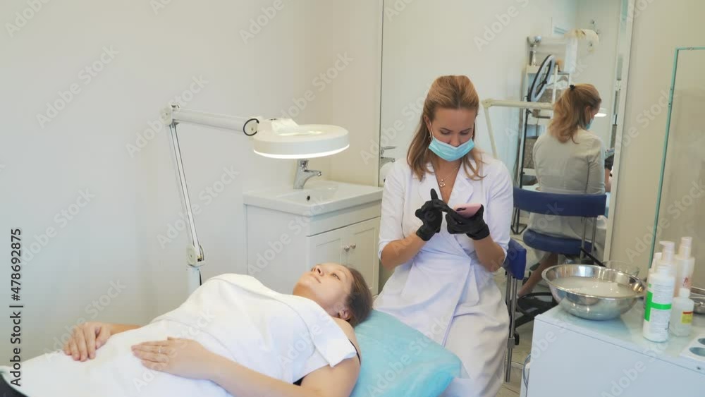 Cosmetologist in white coat is sitting in office and typing text on smartphone. Black gloves on his hands, protective mask on his face. Patient is lying in front of her. Break, online communication