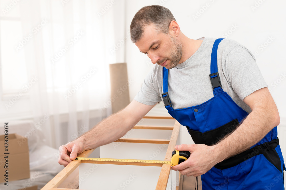 Male Worker In Coverall Measuring Wooden Shelf Assembling Furniture Indoors Male Worker In Coverall Measuring Wooden Shelf Assembling Furniture Indoors