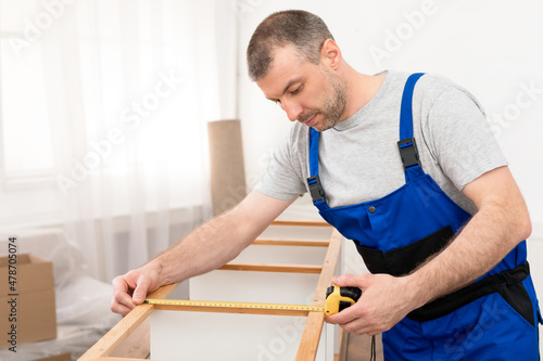 Male Worker In Coverall Measuring Wooden Shelf Assembling Furniture Indoors