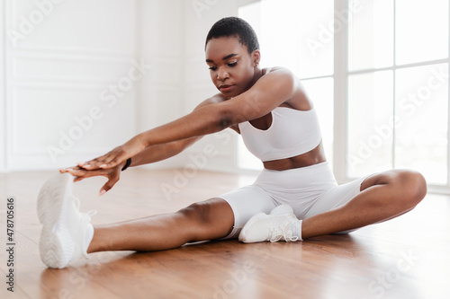 Cheerful Black Woman In White Sportswear Stretching Leg On Floor
