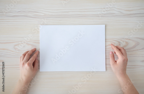 Female hands with white blank paper sheet on wooden table background, top view, empty space, flat lay