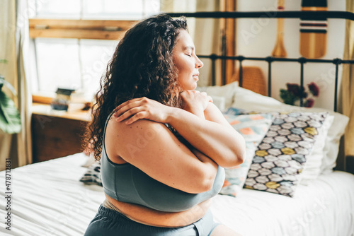 Curly haired overweight young woman in grey top and shorts with satisfaction on face accepts curvy body shape standing in stylish bedroom closeup