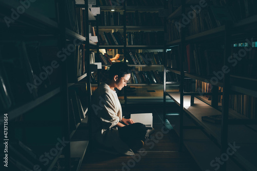 Thoughtful female sitting against bookshelf with a book on the library floor. © Mariia Korneeva
