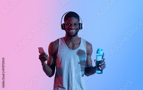 Happy black guy with cellphone and headset picking music playlist for training, holding bottle of water in neon light