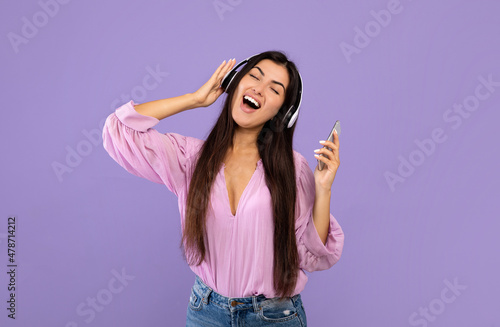 Happy armenian lady with smartphone and wireless headphones, listening to music and singing over purple background