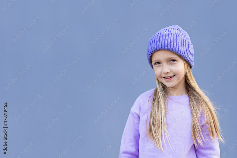 Cute little caucasian girl eight years old with blonde hair smiling outdoor. Kid wearing stylish shirt and knitted hat violet color. Trendy color of the 2022 year very peri Cute little caucasian girl eight years old with blonde hair smiling outdoor. Kid wearing stylish shirt and knitted hat violet color. Trendy color of the 2022 year very peri