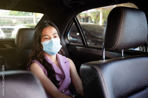 Beautiful Asian businesswoman wear a hygiene protective face mask taking a back seat in taxi portrait, happy Asian woman passenger with face mask traveling by using taxi during the COVID-19 pandemic.