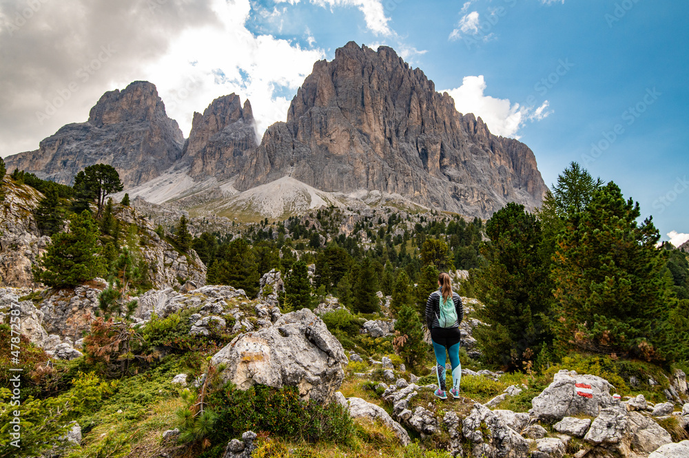 Young woman with turquoise backpack looking at mountain rock towers of Langkofel Group, Grohmannspitze mountain, Fuenffingerspitze mountain and Langkofel Mountain, Sella Pass, Dolomites, Alto Adige
