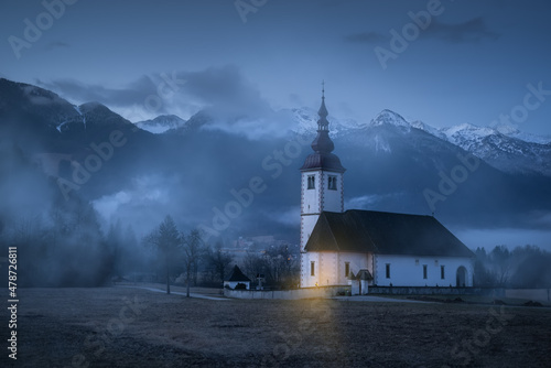 Fotografie, Obraz Mystical and gloomy view of the white church and the chapel standing by the Alpine mountains
