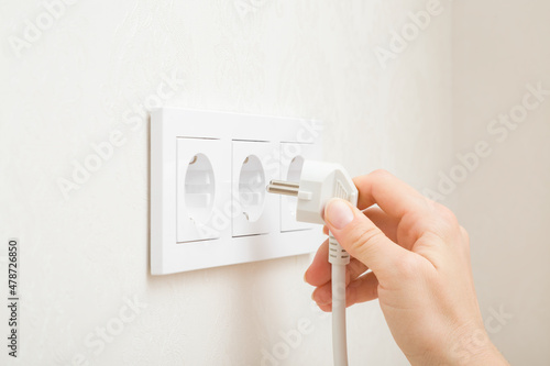 Young adult woman hand holding and plugging white electrical plug in wall outlet socket at home. Closeup. Side view. photo