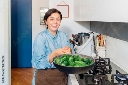 Smiling woman cooking spinach in frying pan at home kitchen