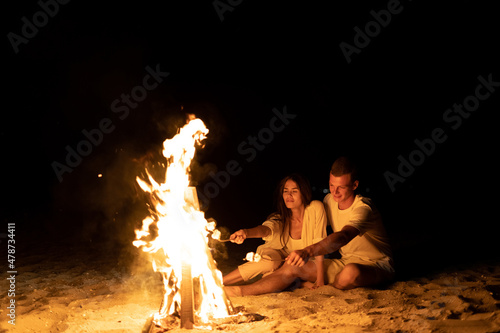 A young couple on the seashore sits by the fire and toasts marshmallows on a stick. A romantic date by the fire. Marshmallow kebab. Tourism and travel concept.