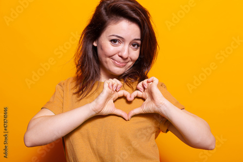Positive adult making heart shape with fingers at camera. Portrait of romantic person doing love sign and symbol with hands, showing affection and emotion over orange background.