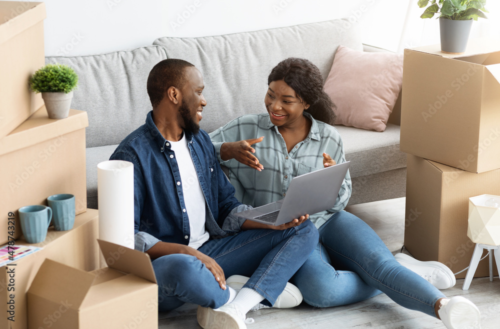 Cheerful Black Married Couple Sitting With Laptop Among Unpacked Cardboard Boxes Cheerful Black Married Couple Sitting With Laptop Among Unpacked Cardboard Boxes