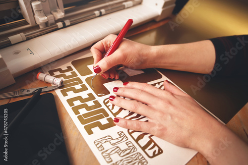 woman with red painted nails removes excess material with weeding tool to make 2022 tiger design stickers from gold vinyl foil. workplace with cutting machine, tools, tablet computer. selective focus