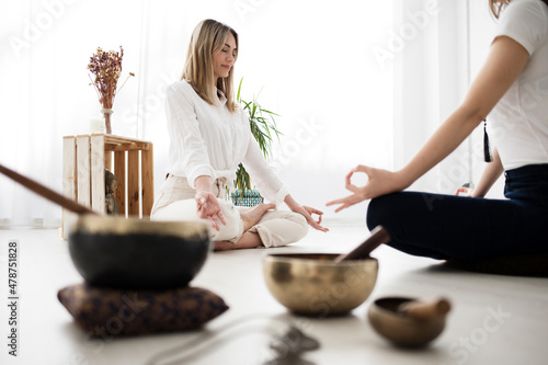 A yoga teacher and her student meditate sitting across from each other. There are various objects such as Tibetan bowls, a Buddha figure and dried flowers.