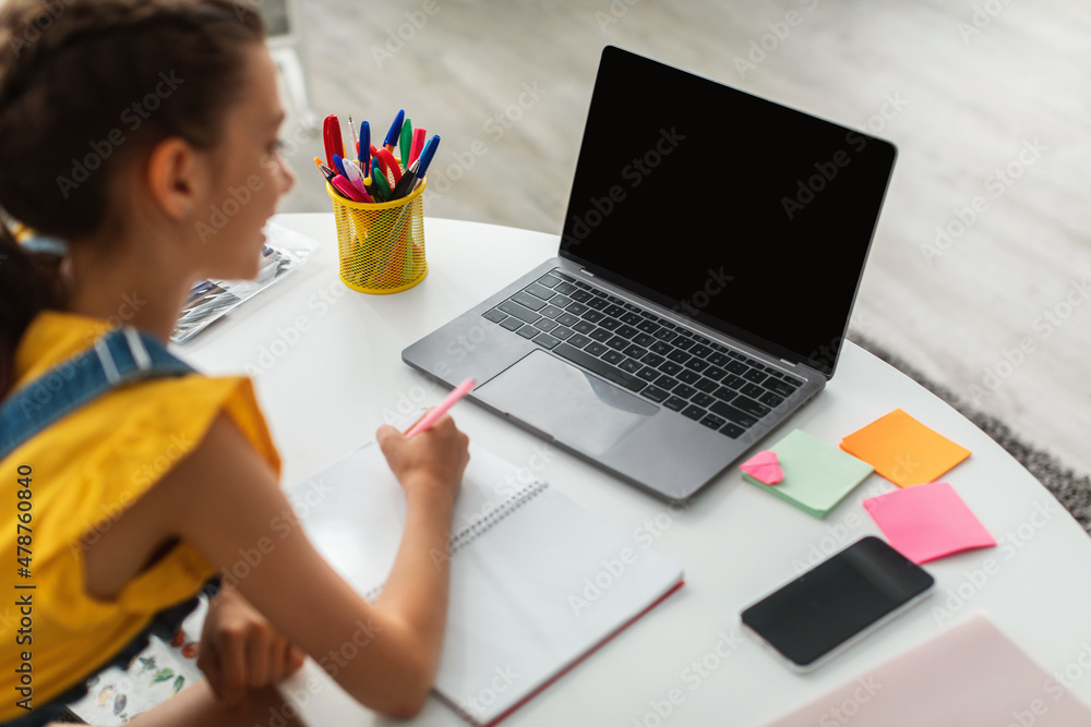 Girl sitting at desk, using blank laptop, writing in textbook Girl sitting at desk, using blank laptop, writing in textbook