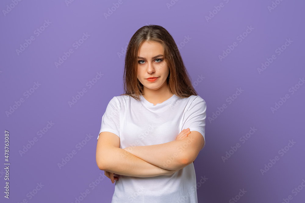 Portrait of upset brunette woman in white casual t shirt standing with arms folded over purple background. Attractive young woman in closed posture Portrait of upset brunette woman in white casual t shirt standing with arms folded over purple background. Attractive young woman in closed posture
