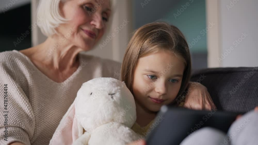 Positive grandmother watching something on tablet with her granddaughter on the couch Positive grandmother watching something on tablet with her granddaughter on the couch