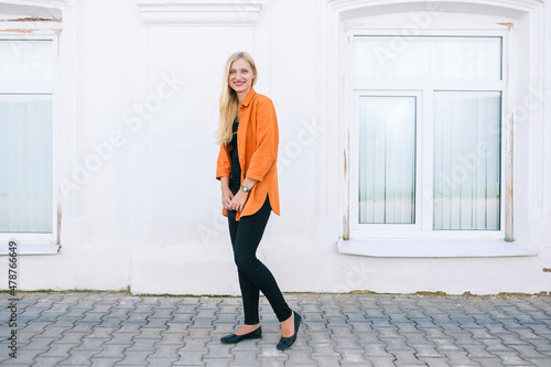 Smiling young girl in an orange shirt and black pants near a white wall. Happy blonde outdoors in an urban environment. Positive and joyful concept with copy space.
