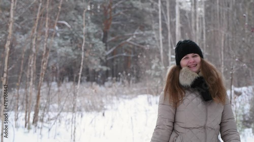 Red-haired girl throws snowballs, smiles and laughs in winter forest in warm clothes: black hat and light jacket, 4k. Blurred forest background
