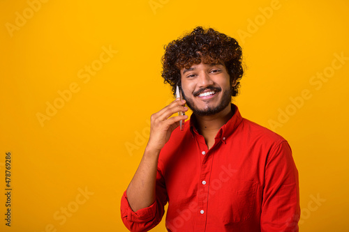 Young adult man with beard talking phone, calling to discuss plans, looking away with dreamy facial expression. Indoor studio shot isolated on orange background © Vadim Pastuh