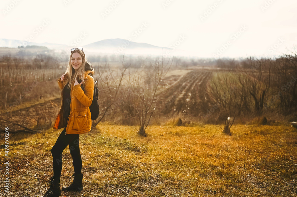 Young happy backpacker woman tourist traveling alone and posing at dawn with fog on background of beautiful nature with mountains in autumn. Blonde smiling lady in yellow down jacket