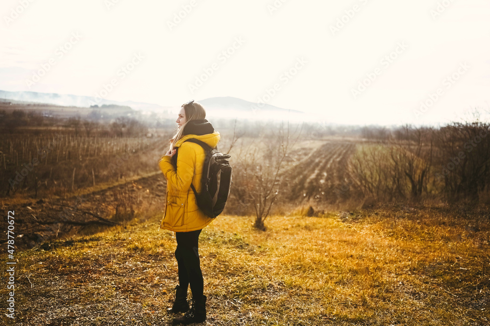 Young happy backpacker woman tourist traveling alone and posing at dawn with fog on background of beautiful nature with mountains in autumn. Blonde smiling lady in yellow down jacket