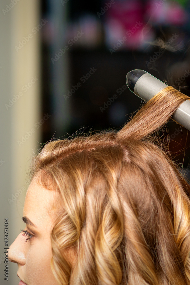 Fototapeta premium Hairdresser makes curls with a curling iron for the young woman with long brown hair in a beauty salon