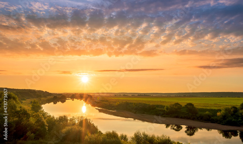 Scenic view at beautiful sunset or sunrise on a shiny river with green bushes on sides  golden sun rays  calm water  deep blue cloudy sky and forest on a background  spring landscape