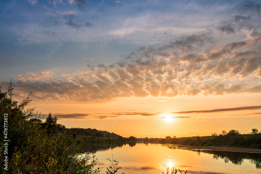 Scenic view at beautiful sunset or sunrise on a shiny river with green bushes on sides, golden sun rays, calm water ,deep blue cloudy sky and forest on a background, spring landscape