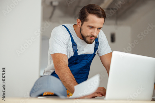 Focused professional handyman in workwear leaning on hands and looking at the screen of a laptop while standing indoors