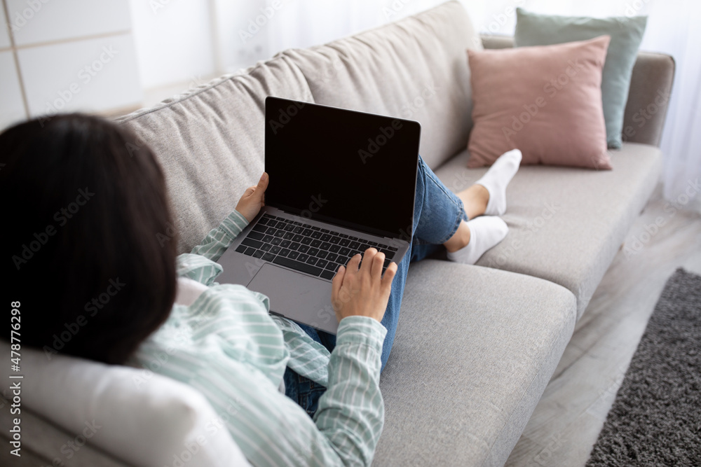 Naklejka premium Unrecognizable Indian woman with laptop lying on comfy sofa and working remotely from home, mockup for website design