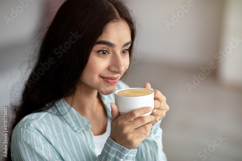 Young Indian woman having coffee break, drinking frothy cappuccino at home, copy space