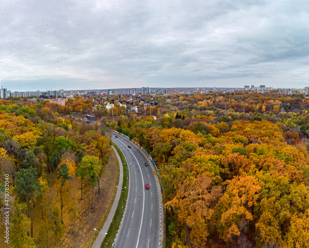Aerial scenic asphalt road curve in autumnal forest near residential district with cloudy sky. Fly above street in autumn city park. Treetop view on Kharkiv, Ukraine
