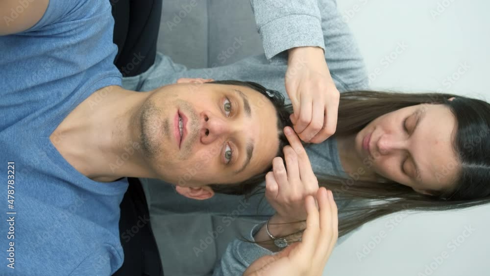 Man blogger is speaking at camera while woman is doing his hair french braids. He is smiling and talking recording his beauty vlog, vertical video. She is sitting on couch. Man blogger is speaking at camera while woman is doing his hair french braids. He is smiling and talking recording his beauty vlog, vertical video. She is sitting on couch.