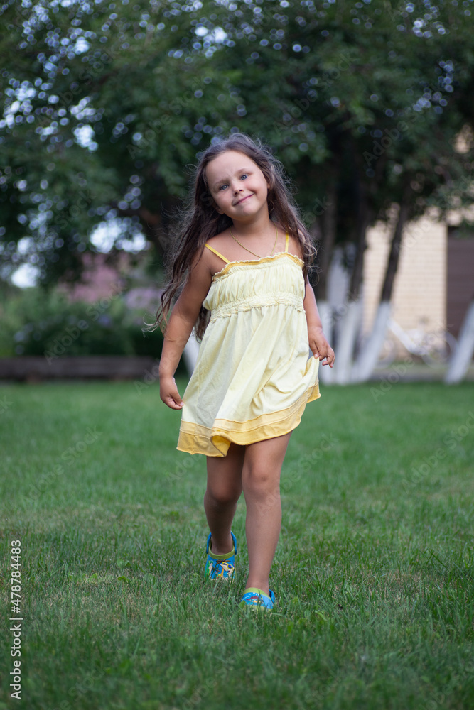 Portrait of little charming smiling girl walking and running around in park wearing light yellow summer dress and slippers in daytime. Adorable background full of green grass and trees.
