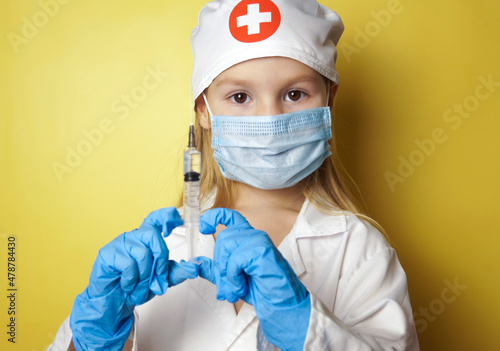 A cute little girl of 5 years old in a doctor's suit with a syringe in her hands on a yellow background. A cheerful child is playing doctor. 