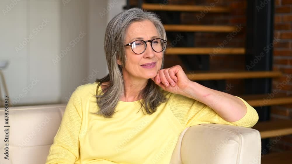 Positive and healthy modern senior woman sitting on the sofa at modern living room, looking aside, then looks at the camera, charming elderly female dreaming. Wellbeing and healthy lifestyle concept Positive and healthy modern senior woman sitting on the sofa at modern living room, looking aside, then looks at the camera, charming elderly female dreaming. Wellbeing and healthy lifestyle concept