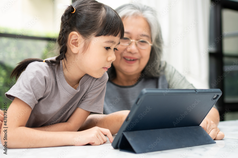 Asian Grandmother with her two grandchildren having fun and playing education games online with a digital tablet at home in the living room. Concept of online education and caring from parents. Asian Grandmother with her two grandchildren having fun and playing education games online with a digital tablet at home in the living room. Concept of online education and caring from parents.