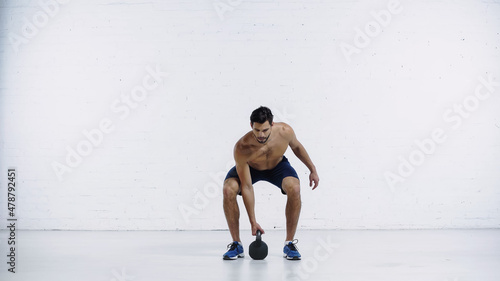 bearded sportsman exercising with heavy kettlebell near white brick wall