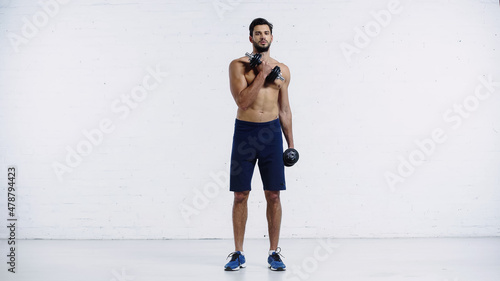 full length of sportive man in shorts and sneakers working out with dumbbells on white