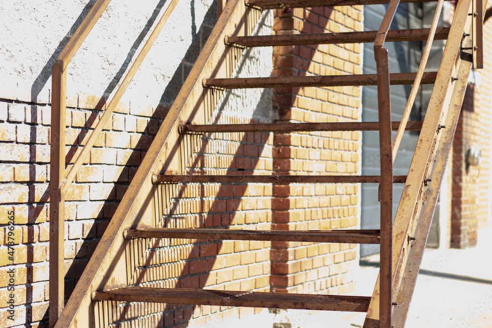 Close up of outdoor metal stairway going to the fire escape. Garage doors on the side of the building. Shadows cast against the brick wal.  