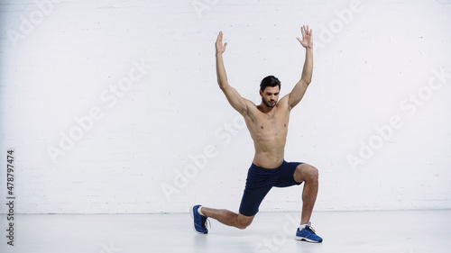 full length of muscular sportsman doing lunges with raised hands near white brick wall