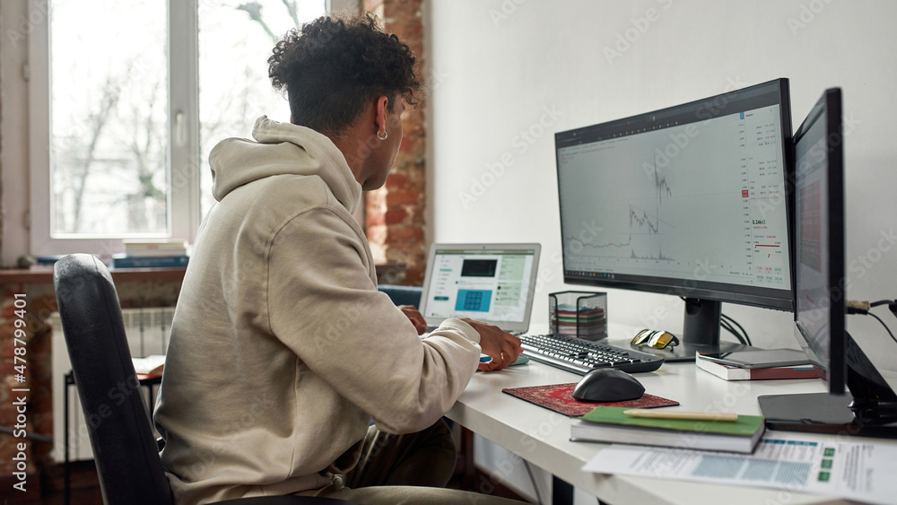 Fototapeta premium Side view of young guy trading online using pc and laptop, sitting at home in modern living room office