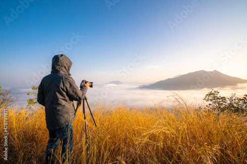 professional photographer taking a photo with camera lens and tripod on top of mountain with meadow and cloud fog landscape with beautiful sunrise in autumn before summer