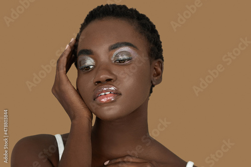 Closeup portrait of a serene young black woman with short Afro hair, light makeup and lipstick posing by herself inside a studio with a pecan background resting her palm of her hand on her face.