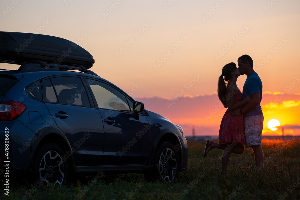 Happy couple spending time together near their SUV car during honeymoon road trip at warm summer evening. Young man and woman enjoying road trip travelling by vehicle in nature.