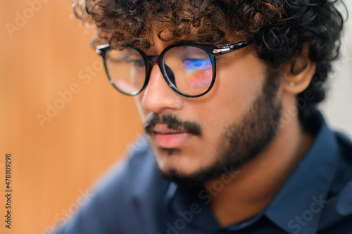 Selective focus at the reflection of screen on the glasses of Indian male employee, concentrated multi ethnic male freelancer in glasses using laptop, indian latin curly developer guy coding