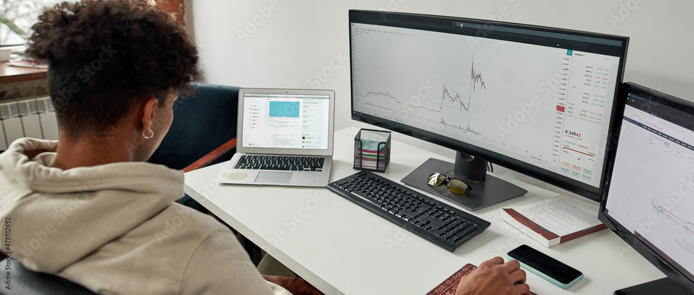Rear view of young male trader using laptop and pc, looking at computer screen to analyze statistics while sitting at home office desk